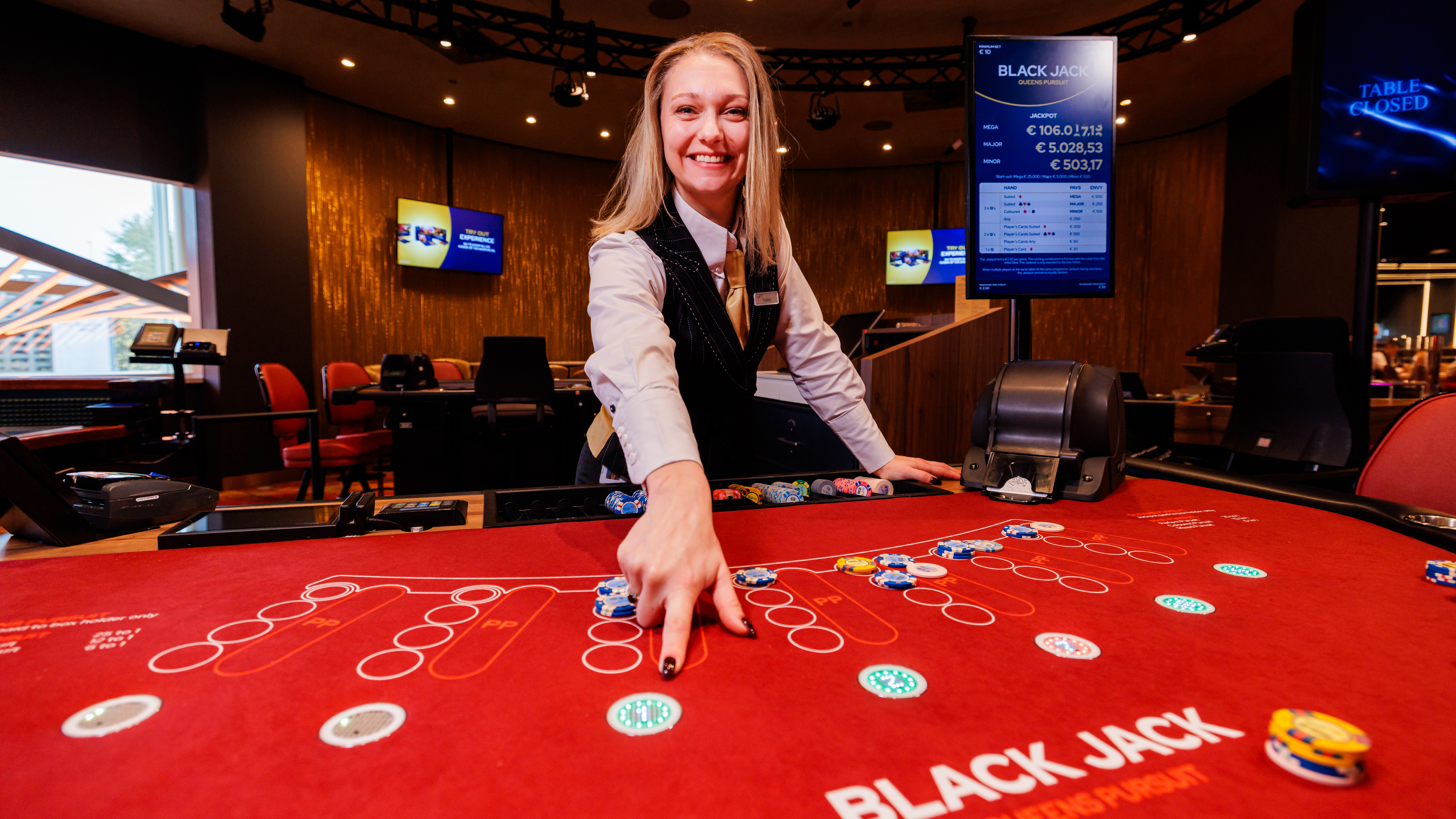 De dealer wijst op de coinspot van de Black Jack "Queens Pursuit" speeltafel